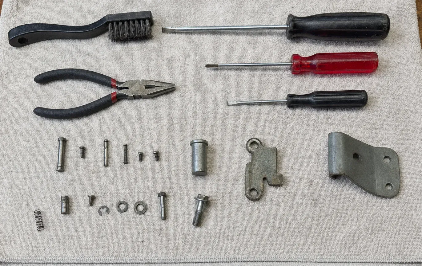 A hinge plate, bracket, springs, washers and screws laid out on a pale towel with a wire brush, pliers and three screwdrivers